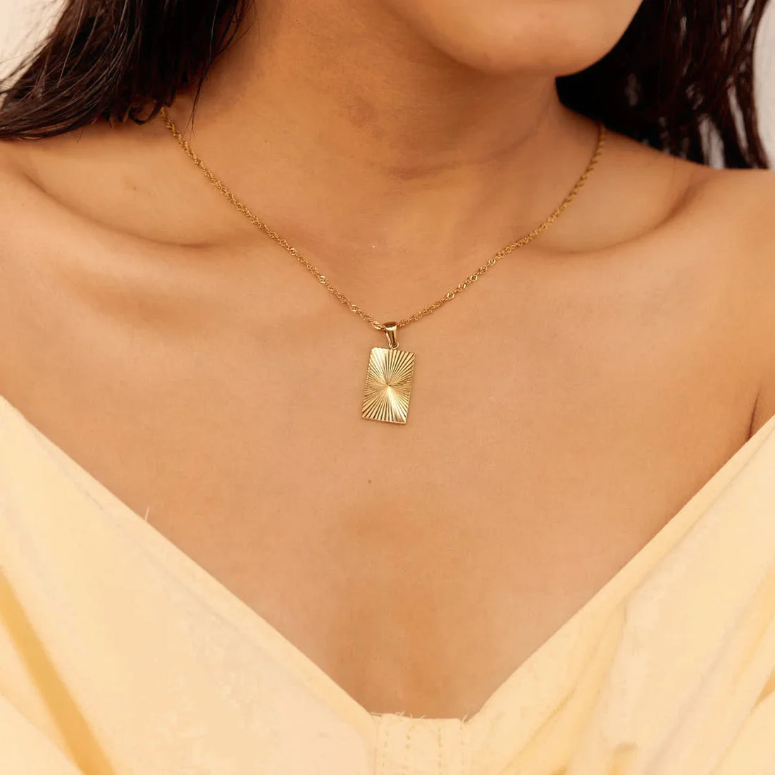 Gold necklace with a rectangular pendant worn by a person in a yellow top.